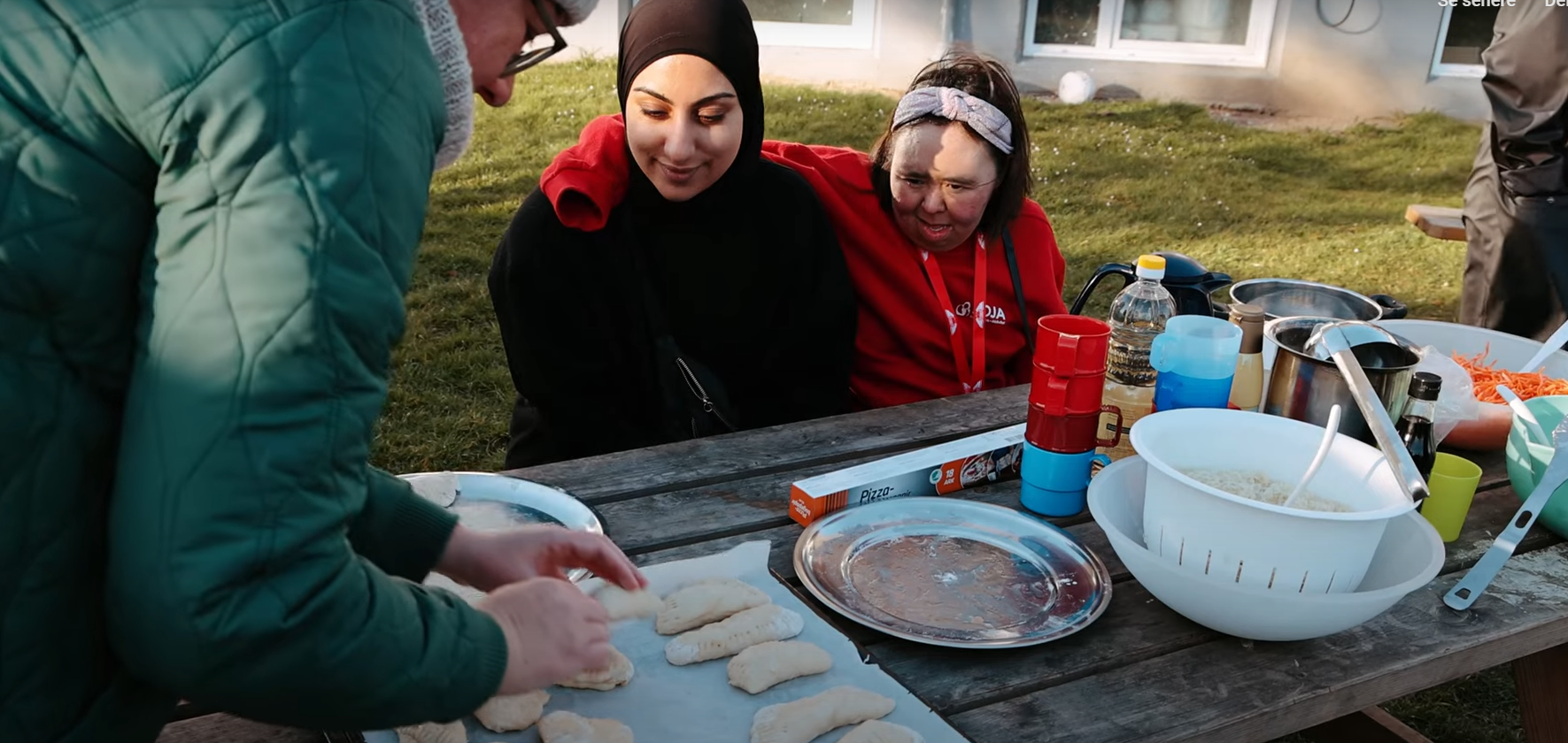 To personer sidder udendørs ved et bordebænke sæt. En tredje person står og lægger brød på en bradepange.