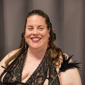 Ung kvinde med brunt hår, lys hud og rød læbestift sidder og smiler i en sort natkjole med blonder, og i hånden har hun pind med sorte fjer på.