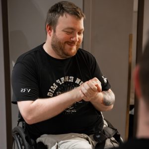 Ung mand med tatoveringer, lys hud, rødligt hår og skæg sidder i hans kørestol og kigger ned i gulvet og smiler, mens han holder sig selv hænderne.