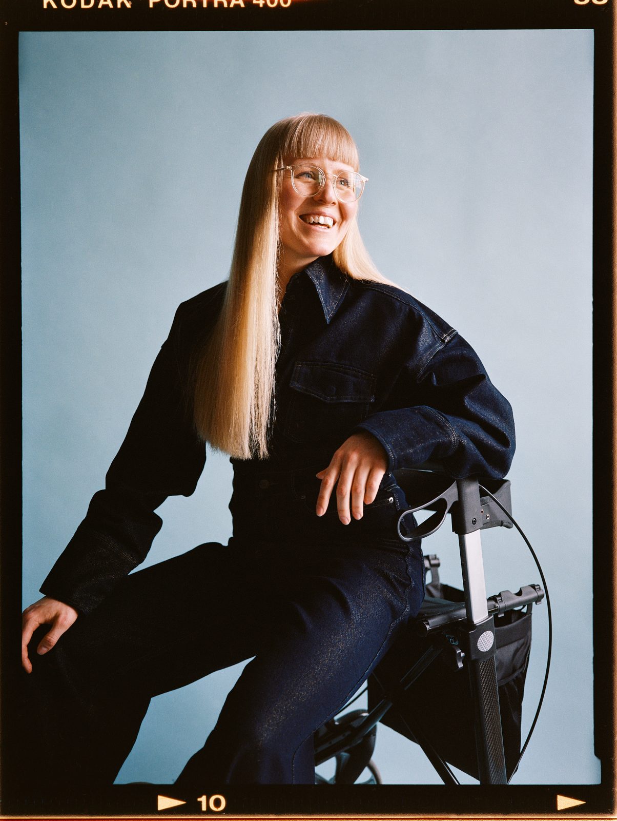 Portræt af ung kvinde med langt lyst hår og briller. Hun sidder på en rollator og smiler.