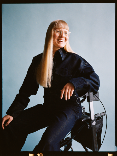 Portræt af ung kvinde med langt lyst hår og briller. Hun sidder på en rollator og smiler.